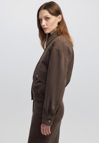 Bruine, taille-lange jas met een kraaghalslijn, elastische taille, grote vo pockets en drukknoopdetails; gladde textuur.