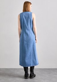 Vaaleansininen farkkumekko, jossa on istuva siluetti, hihaton malli, pitkä pituus ja näkyvät saumat. Mustat polvisukat paksuilla pohjilla.