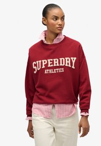 Sudadera roja con "SUPERDRY ATHLETICS" en texto crema, superpuesta a una camiseta de rayas en rosa claro y blanco. Pantalones blancos, de textura suave.