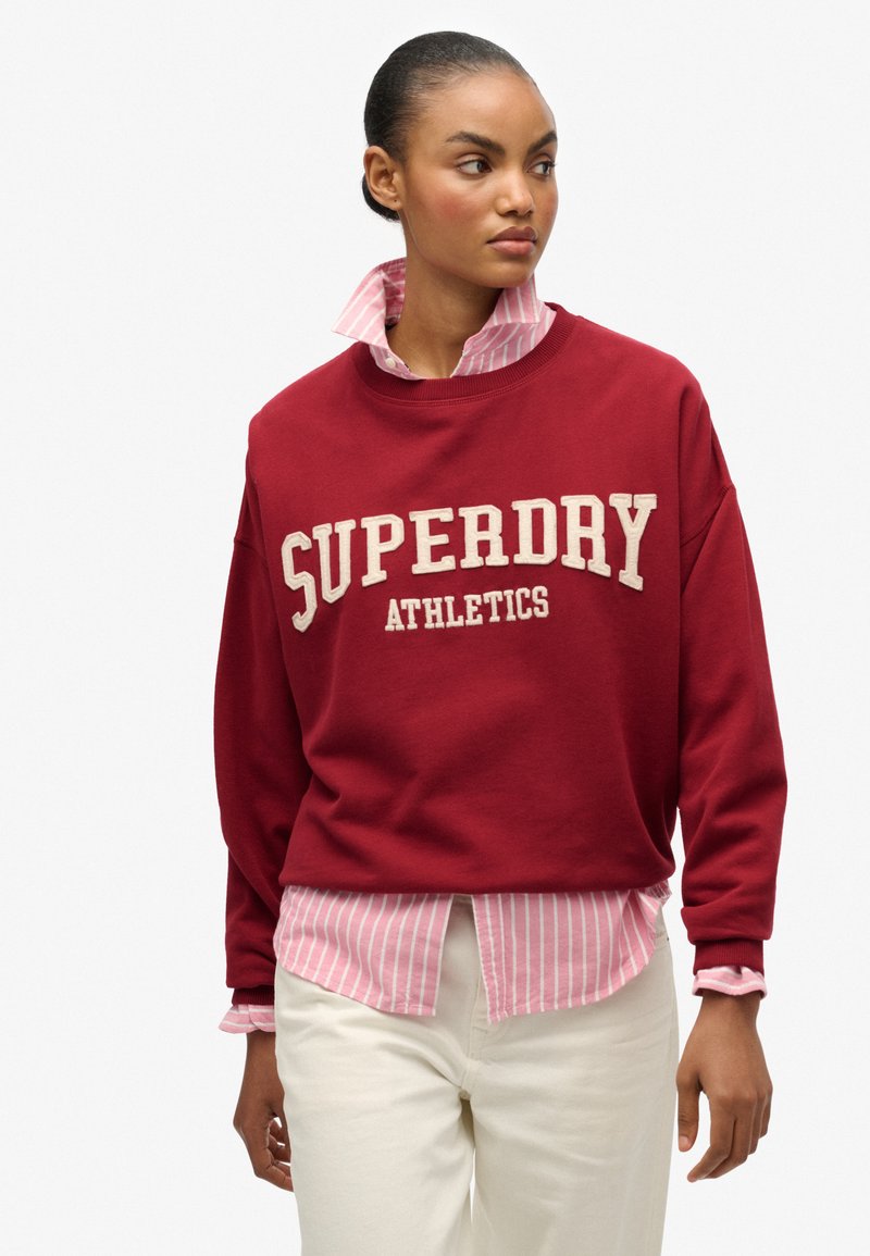 Sudadera roja con "SUPERDRY ATHLETICS" en texto crema, superpuesta a una camiseta de rayas en rosa claro y blanco. Pantalones blancos, de textura suave.