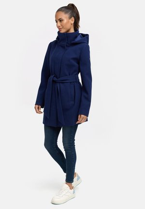 Navy blue, belted hooded coat with a high collar, made of soft fabric. Features side pockets and long sleeves. Paired with dark jeans and sneakers.