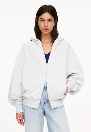 Sweat à zip gris clair avec une capuche spacieuse, des épaules tombantes et des poignets côtelés. Fait en tissu doux, porté sur un haut bleu marine et associé à un jean clair.