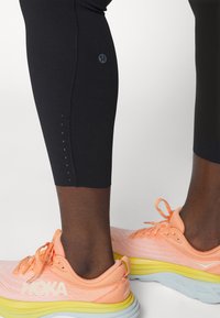 Leggings atléticos negros con detalles reflectantes cerca del dobladillo, acompañados de zapatillas de correr en naranja y amarillo con una parte superior de malla texturizada.