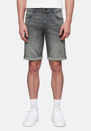 Mand iført falmede grå cowboyshorts med opkrogede kanter, kombineret med hvide sokker og hvide sneakers, stående mod en ensfarvet baggrund.