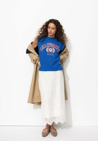 T-shirt sans manches bleu avec impression "CALIFORNIA", associé à une jupe en dentelle blanche et un trench-coat beige, accessoirisé avec des ballerines à motif léopard.