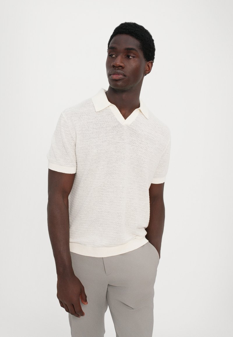Polo-shirt en lin beige clair texturé à manches courtes avec un col en V et des finitions crème, associé à un pantalon ajusté gris. Design décontracté et respirant.