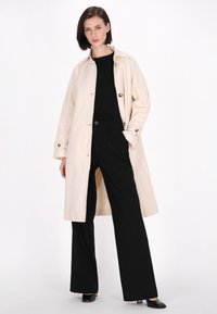 Beige trenchcoat med løs pasform og store knapper, parret med sorte brede bukser og sorte højhælede sko. Glat stoftekstur.