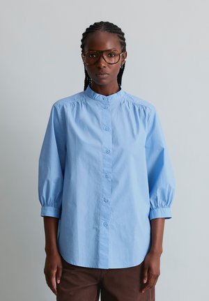 Vrouw met gevlochten haar die grote bruine bril draagt, een lichtblauw overhemd met driekwartmouwen en een bruine broek, staand tegen een effen achtergrond.