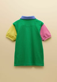 Polo vert à manches courtes avec un col bleu, une manche gauche jaune et une manche droite rose, posé à plat sur un fond beige.