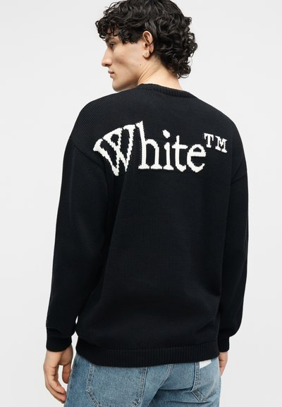 Νεαρός άνδρας με σγουρά μαλλιά φορώντας μαύρο πουλόβερ με μεγάλο λευκό κείμενο "White™" στο πίσω μέρος, συνδυασμένο με μπλε τζιν, κοιτάζοντας πλαγίως.