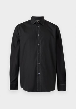Homme portant une chemise noire à boutons, pantalon gris foncé, chaussures habillées noires, tenant un sac à épaule noir sur un fond clair uni.