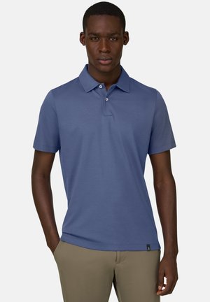 Mann trägt ein blaues Polohemd mit kurzen Ärmeln und beige Hose, steht mit einer Hand in der Tasche vor einem einfarbigen Hintergrund.