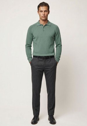 Mand står med hænderne i lommerne iført en grøn langærmet polo, mørkegrå bukser, sort bælte og sorte pæne sko.