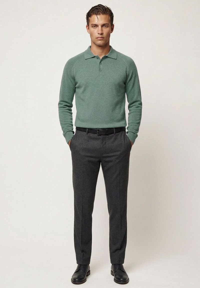 Uomo in piedi con le mani in tasca, indossa una polo verde a maniche lunghe, pantaloni grigio scuro, cintura nera e scarpe eleganti nere.