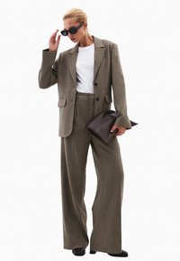 Hnědý kostkovaný blazer se dvěma knoflíky, v kombinaci s kalhotami s vysokým pasem a širokými nohavicemi. Modelka drží tmavě hnědou kabelku a nosí černé sluneční brýle.