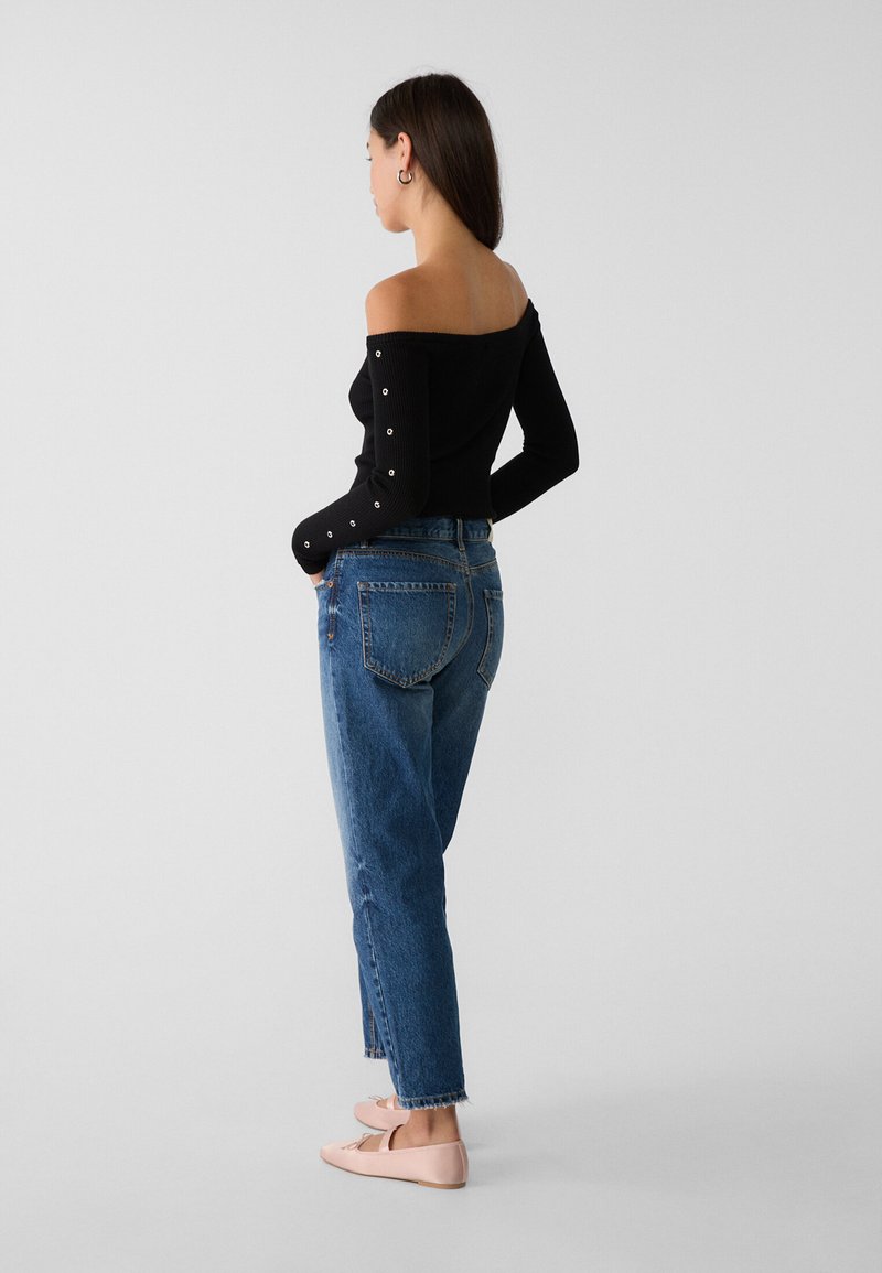 Top a spalle scoperte nero in maglia con maniche a bottoni argentati, abbinato a jeans di denim blu a vita alta e ballerine rosa chiaro.
