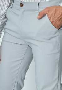 Pantalon gris clair avec une coupe slim, doté d'une fermeture à bouton, de poches latérales et d'une texture de tissu lisse.