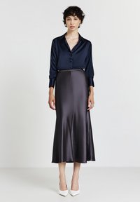 Donna con capelli corti e ricci indossa una blusa di satin navy, una gonna midi di seta grigia e tacchi a punta bianchi, in piedi contro uno sfondo chiaro e semplice.
