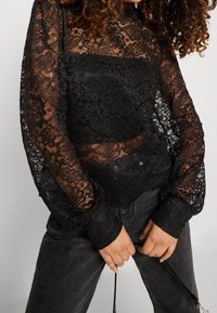 Haut en dentelle noire à manches longues, avec des motifs floraux et des sections transparentes. Associé à un jean foncé, présentant une coupe ajustée.