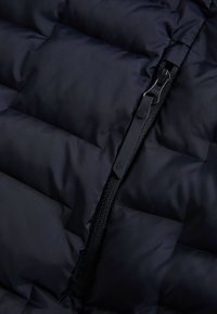 Crna puffer jakna s kvadratnim ušivenim dijelovima, s čvrstim patentnim zatvaračem i glatkom teksturom tkanine. Patentni zatvarač se nalazi sprijeda.