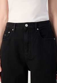 Personne portant un jean en denim noir taille haute avec un bouton et une fermeture éclair, associé à un haut blanc sans manches rentré.