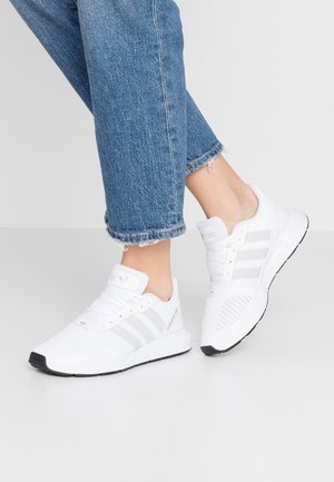 Personne portant des baskets blanches Adidas avec des rayures grises et un jean bleu avec des ourlets effilochés, debout sur un sol gris clair.