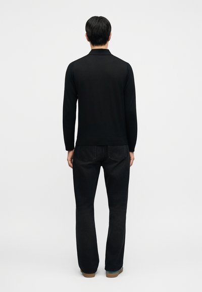 Μαύρο μακρυμάνικο πόλο με γιακά, φορεμένο με σκούρο τζιν. Το ύφασμα φαίνεται λείο και η εφαρμογή είναι χαλαρή. Τα παπούτσια είναι ανοιχτό καφέ.