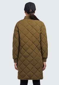 Manteau long matelassé vert olive avec un motif en diamant, des poignets côtelés et un col au dos. Porté avec une casquette noire, vu de l'arrière.