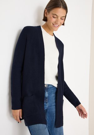 Marineblauer Strickcardigan mit offener Vorderseite, gerippter Textur und aufgesetzten Taschen. Tragen über einem hellen Cremesweater und Jeans.