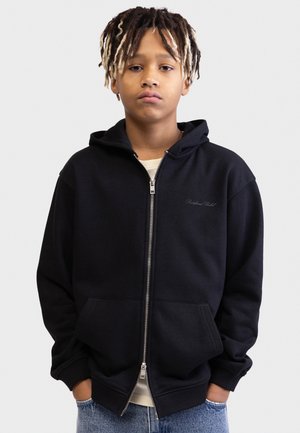 Fiatal személy rövid dreadlock frizurával, fekete cipzáras kapucnis pulóvert és kék farmert visel, sima világos háttér előtt áll.