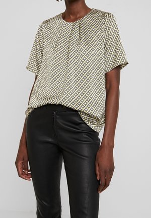 Kobieta ubrana w beżową koszulę w kratkę z krótkim rękawem, wpuszczoną w czarne, wysokiej talii skórzane spodnie, na tle jednolitego tła.