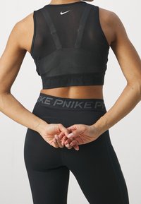 Musta urheilusukka, jossa on verkko-osio, säädettävät hihnat ja leveä elastinen vyö, jossa on "NIKE"-brändäys, yhdistettynä korkeavyötäröisiin mustiin leggings-housuihin.