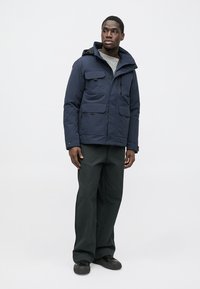 Granatowa ocieplana kurtka z przednimi kieszeniami, regulowanym kapturem i zamkiem błyskawicznym. Noszona z ciemnozielonymi spodniami i czarnymi butami.