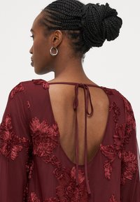 Tumši sarkana kleita ar ziedu izšuvumu, atvērtu muguras dizainu ar saites aizdari un plūstošiem piedurknēm, izgatavota no viegla auduma.