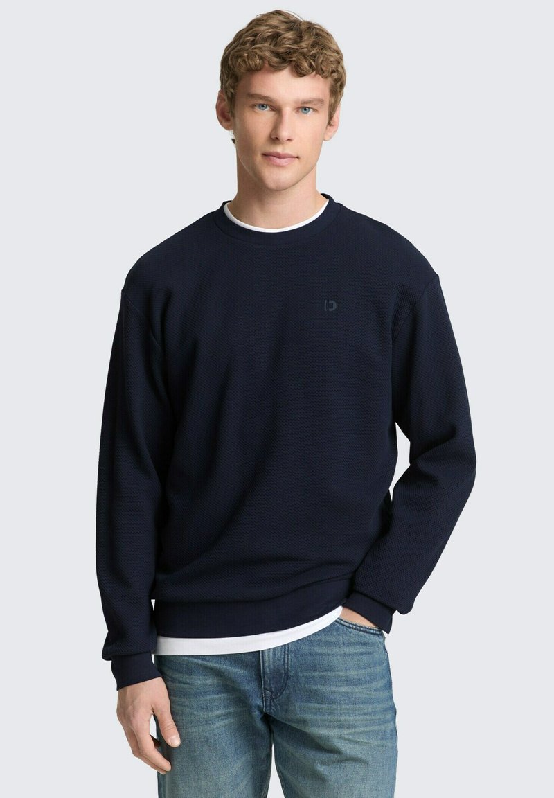 Joven con cabello rizado, vistiendo un suéter azul marino sobre una camisa blanca y jeans azul claro, de pie con una mano en el bolsillo.