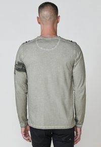 Hosszú ujjú ing tompa zöld anyagból; kerek nyakkal, texturált részletekkel és grafikus díszítéssel a bal ujján.