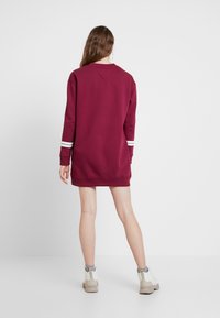 Viininpunainen college-mekko pitkillä hihoilla, jossa on valkoiset raidat hihojen suissa, ribbineckline ja rento malli.