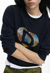 Granatowa bluza z kolorową, abstrakcyjną haftowaną grafiką, ściągaczami na mankietach i okrągłym dekoltem, noszona na białej koszuli.