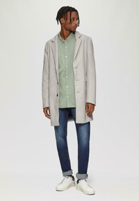 Lichtgrijze wollen jas met gesneden kleppen, enkelvoudige sluiting, gedragen over een groene button-up shirt, jeans en witte sneakers.
