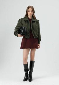 Veste bomber courte vert olive avec manches bouffantes, associée à une robe bordeaux, des bottes noires hautes jusqu'aux genoux, et un petit sac à main noir.