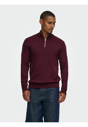 Mand iført en burgunderfarvet langærmet sweater med lynlås foran, kombineret med blå denimjeans, der kigger lidt ned mod sin venstre side.