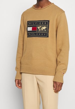 Mężczyzna noszący wielbłądzi sweter dziany z czarno-czerwonym naszywką z logo Tommy Hilfiger i beżowe spodnie na prostym białym tle.