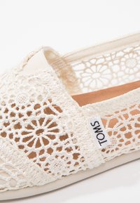 Chaussure blanche à enfiler en dentelle avec des motifs floraux complexes, des accents élastiques et une doublure intérieure beige. Présente une petite étiquette avec le logo TOMS.