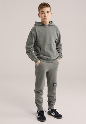 Dječak stoji sa rukama u džepovima noseći sivu hoodicu, odgovarajuće trenerske hlače s aplikacijama i crne tenisice s bijelim prugama.