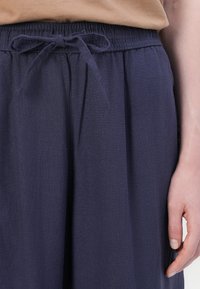 Navyblauwe broek met een trekkoord in de taille, gemaakt van zachte stof. Heeft plooien en een relaxte pasvorm. Hand zichtbaar aan de zijkant.