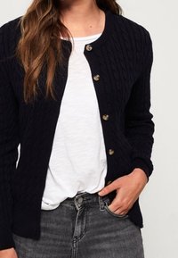 Marineblaue Strickjacke mit Kabelmuster und Rundhalsausschnitt, ausgestattet mit goldenen Knöpfen und gerippten Bündchen. Tragen über einem weißen Hemd und grauen Jeans.