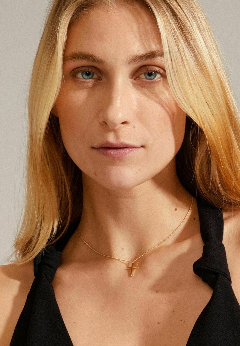 Collana d'oro con pendente a croce, indossata sulla pelle nuda. Lo sfondo presenta una tonalità neutra, mettendo in risalto i dettagli e la lucentezza della collana.