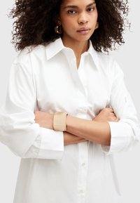 Donna con capelli ricci che indossa una camicia bianca con bottoni, grandi orecchini geometrici dorati e un ampio bracciale beige, con le braccia incrociate.
