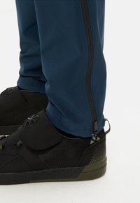Blauwe sportbroek met een ritsdetail aan de zijkant, gecombineerd met zwartgroene schoenen met een gestructureerd oppervlak en stevige sluitingen.