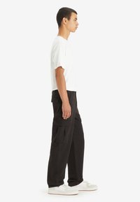 Sorte cargo-bukser med flere sidelommer, lavet af slidstærkt stof, sammen med en ensfarvet hvid t-shirt og hvide sneakers. Sidevisning.
